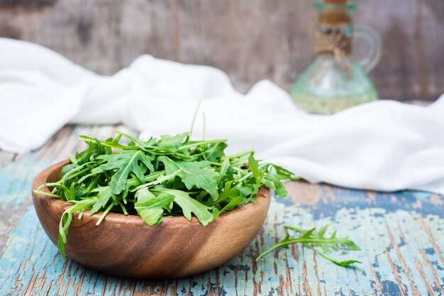 Frische Blätter von Arugula in einer hölzernen Schüssel, in einem Krug mit Öl und in einer Serviette auf einem Holztisch