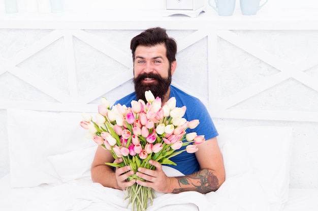 Frische Blumen überraschen Geschenk für Ehepartner Bärtiger Hipster im Bett Frühlingsstimmung in seinem Schlafzimmer Mann hält Tulpenstrauß beim Entspannen im Bett Blumenlieferdienst Geburtstag Jahrestag Urlaub