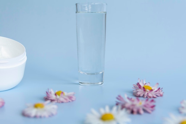 Frische Blumengänseblümchen werden in einer Ecke mit einem Glas sauberem Wasser und einem weißen Glas mit einer cremefarbenen Kosmetik auf einem blauen Hintergrund ausgelegt