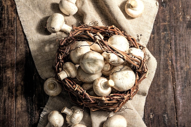 Frische Champignons in einem Korb. Rohzutat zum Kochen veganer Speisen. Alter hölzerner Hintergrund, hartes Licht der Mode, dunkler Schatten, rustikaler Stil, Draufsicht