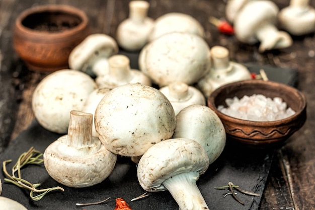 Frische Champignons mit Gewürzen und Kräutern. Rohzutat zum Kochen veganer Speisen. Alter hölzerner Hintergrund, hartes Licht der Mode, dunkler Schatten, rustikaler Stil, Nahaufnahme