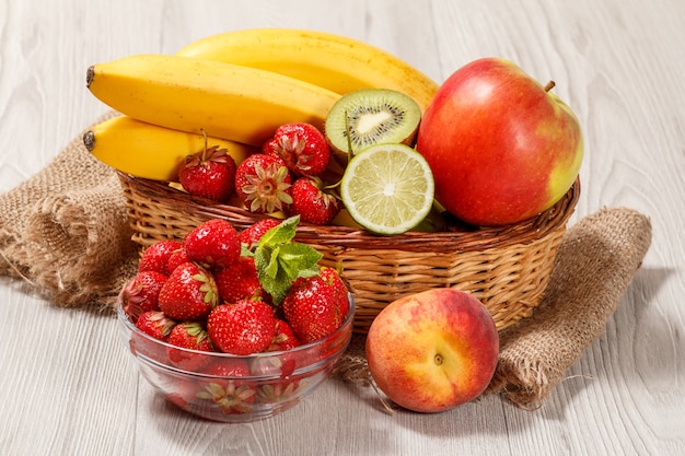 Frische erdbeeren in der glasschüssel mit minzblättern, nektarine, limette, kiwi, banane, apfel in einem weidenkorb auf einem holztisch