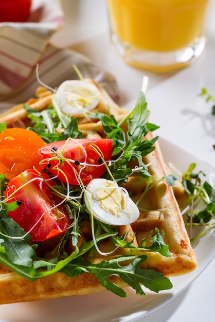 Frische gebackene süße belgische Waffeln mit Eiern, Tomaten, Mikrogrün und Avocado dienten auf einer Platte mit Orangensaft auf Marmor