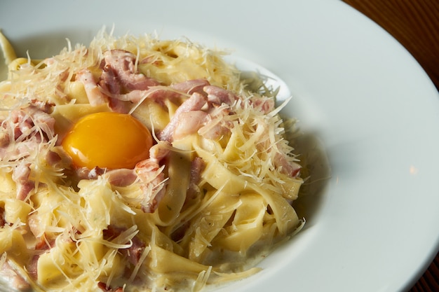 Frische hausgemachte italienische Carbonara-Nudeln mit rohem Eigelb, Parmesan, weißer Soße, Speck in der weißen Schüssel auf hölzernem Hintergrund.