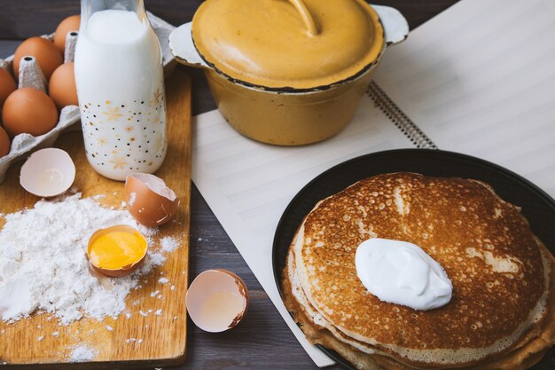 Frische, heiße Pfannkuchen in einer Pfanne, Eier, Milch, Mehl auf einem Holztisch. Ansicht von oben