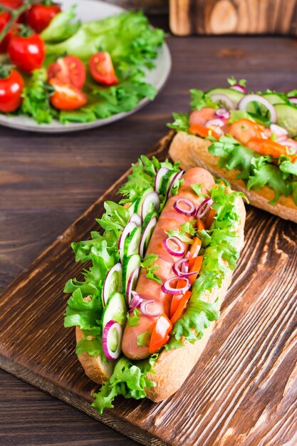 Frische Hot Dogs mit Wurst, Gurken, Tomaten und Kräutern auf einem Holztisch Fast Food zu Hause Vertikale Ansicht Nahaufnahme