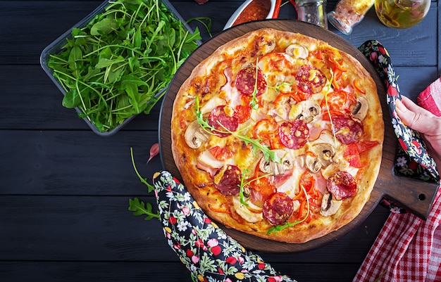 Frische italienische Pizza mit Hühnerleiste, Pilzen, Schinken, Salami, Tomaten, Käse auf einem schwarzen Hintergrund in den Händen.