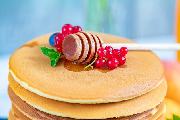 Frische köstliche Pfannkuchen mit Sommerhimbeeren, Blaubeeren, roten Johannisbeeren, Aprikose, Pfirsich, Minze und Honigschöpflöffel in Nahaufnahme. Süßes, nahrhaftes Essen. Geringe Schärfentiefe