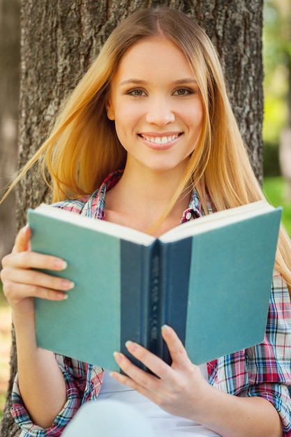 Frische Luft und Lieblingsbuch. Schöne junge Frau, die ein Buch hält und lächelt, während sie sich an den Baum in einem Park lehnt