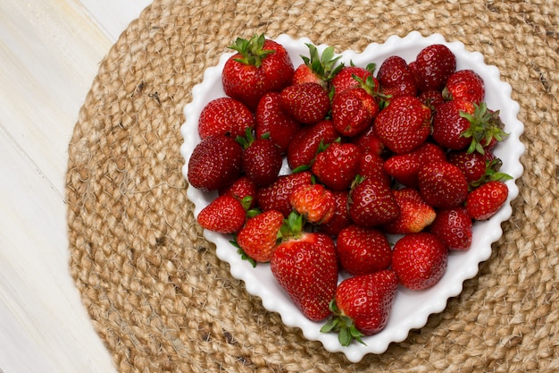 FRISCHE REIFE ERDBEEREN AUF EINER HERZWEISSEN PLATTE MIT EINEM JUTE-TISCH