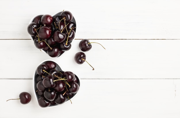 Frische reife rote und schwarze Kirschen in der zwei Herz-förmigen Schüssel auf weißem hölzernem Plankenboden.