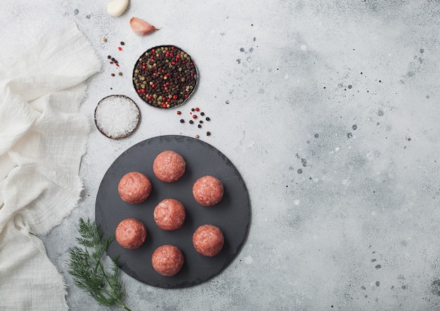 Frische rohe Hackfleischbällchen auf rundem Steinbrett mit Pfeffer, Salz und Knoblauch auf heller Tischoberfläche mit Dill und Handtuch. Platz für Text