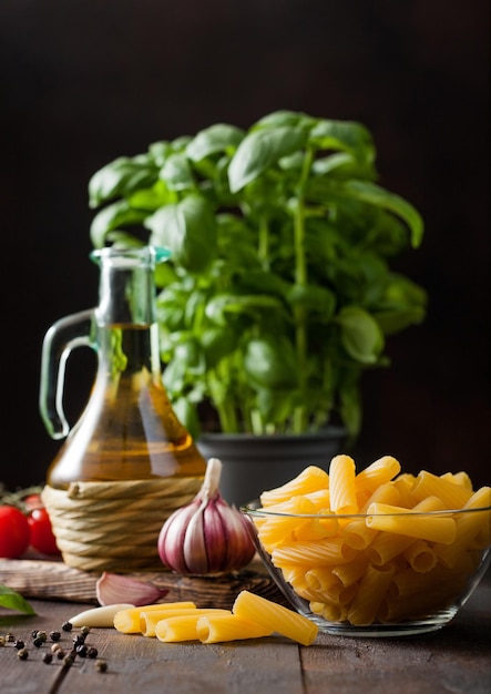 Frische rohe Penne-Nudeln in Glasschüssel mit Basilikum-Pflanzenöl und Tomaten mit Knoblauch und Pfeffer auf Holztischhintergrund