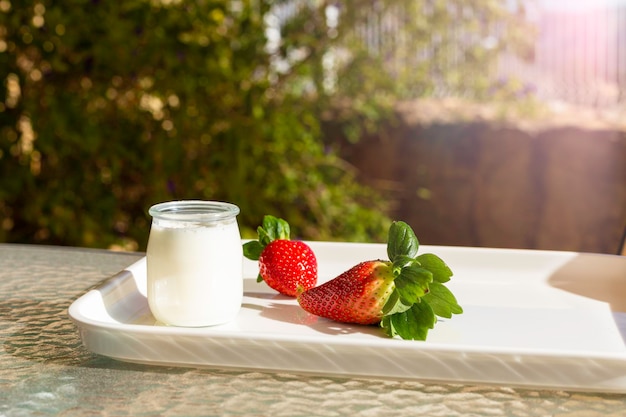 Frische rote Erdbeeren und weißer Joghurt in einem Glasgefäß auf einem weißen Tablett auf einem Tisch im Garten oder auf der Terrasse an einem sonnigen Morgen