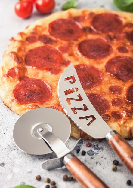 Foto frische runde gebackene italienische pepperoni-pizza mit radschneider und messer mit tomaten und basilikum auf hellem küchentischhintergrund.