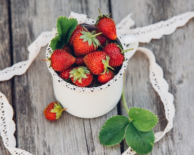 Frische saftige Erdbeeren mit Blättern. Rustikaler emaillierter Becher und handgemachte Spitze. Retro-Magazinbild.