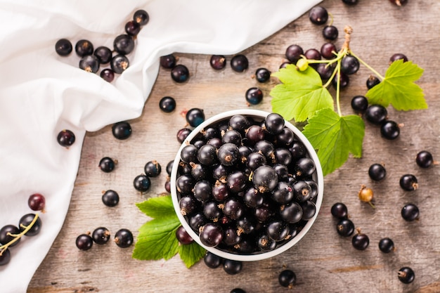 Frische Schwarze Johannisbeere in der Schüssel. Viele Johannisbeeren und Blätter verstreuten sich auf dem schäbigen Tisch und dem weißen Tuch. Ansicht von oben