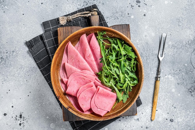 Frische Schweineschinkenscheiben in Holzplatte mit Rucolasalat Grauer Hintergrund Ansicht von oben