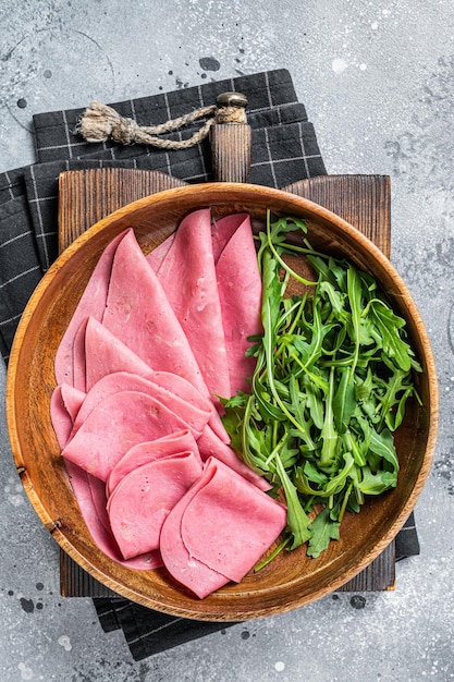 Frische Schweineschinkenscheiben in Holzplatte mit Rucolasalat Grauer Hintergrund Ansicht von oben