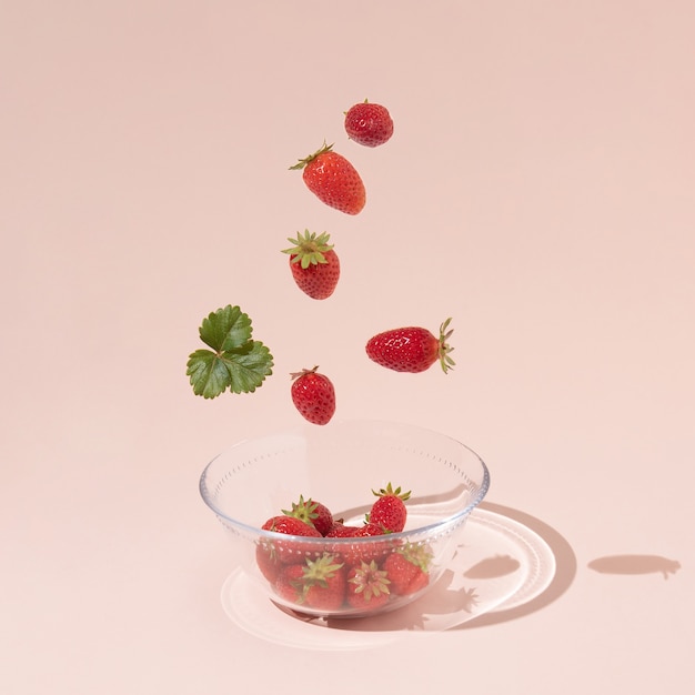 Frische Sommerfrüchte, Erdbeeren und grüne Blätter fallen in eine Glasschüssel, einzeln auf rosafarbenem Hintergrund. Kreatives Essenslayout. Quadrat