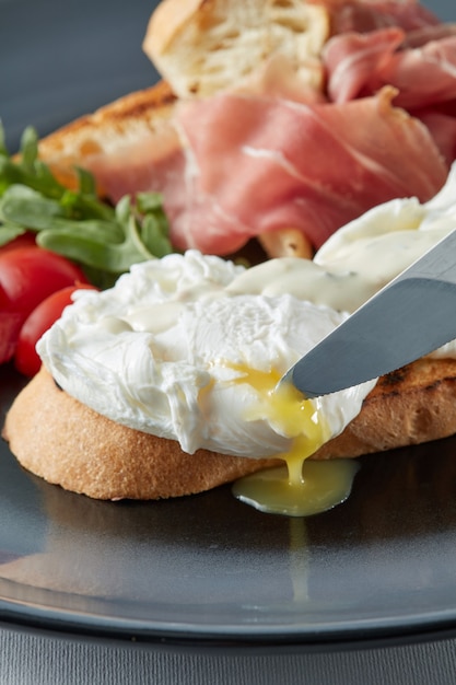 Frische Toastsandwiches mit pochiertem Ei, Tomate, Salat und Speck auf Teller, Messer, das ein Ei schneidet