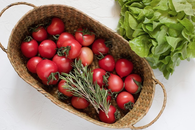 Frische Tomaten einige Zweige Rosmarin in einem Weidenkorb mit Salatblättern auf einer weißen Oberfläche