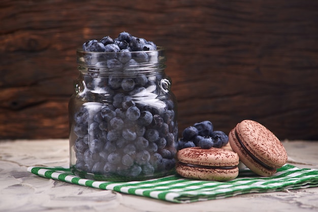 Frische und geschmackvolle Blaubeerbeeren in einem Glasgefäß auf einer karierten Serviette mit süßen Makronen