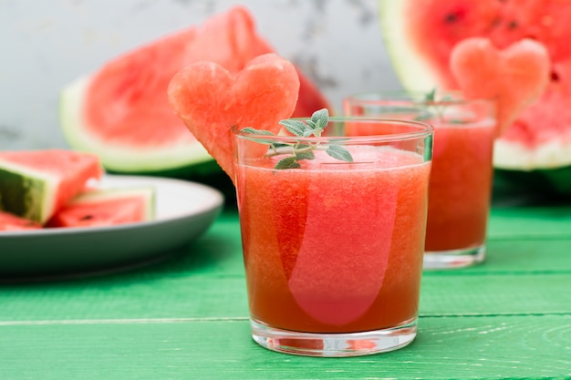 Frische Wassermelone mischte Getränk mit tadellosen Blättern und einem Herzen der Wassermelone in den Gläsern und in den Scheiben der Wassermelone auf einem Holztisch