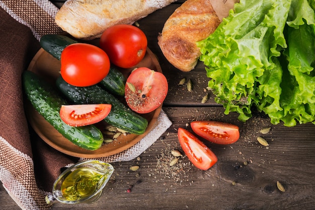 Frische Zutaten zum Kochen von Salat: Tomate, Gurke, Salat, Olivenöl und Gewürze über rustikalem Holztischhintergrund. Serviert mit frischem Baguette. Konzept für gesunde Ernährung