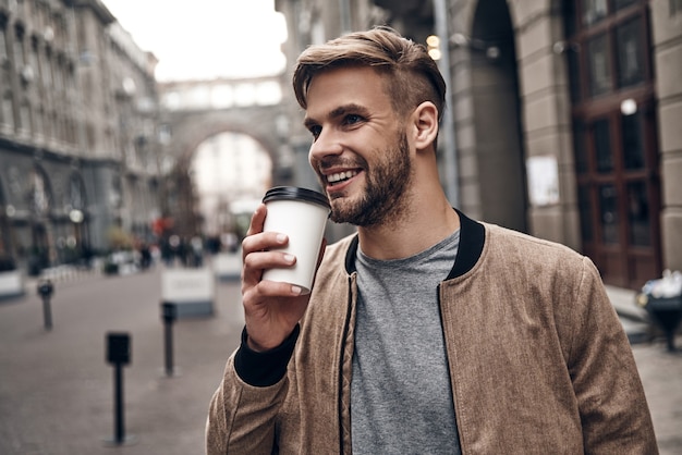 Frischen Kaffee genießen. Hübscher junger Mann in Freizeitkleidung, der Einwegbecher hält und lächelt, während er durch die Stadtstraße geht walking