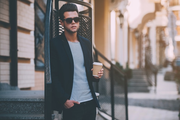 Frischen Kaffee genießen. Schöner junger Mann in eleganter Freizeitkleidung, der eine Kaffeetasse hält, während er auf der Straße steht