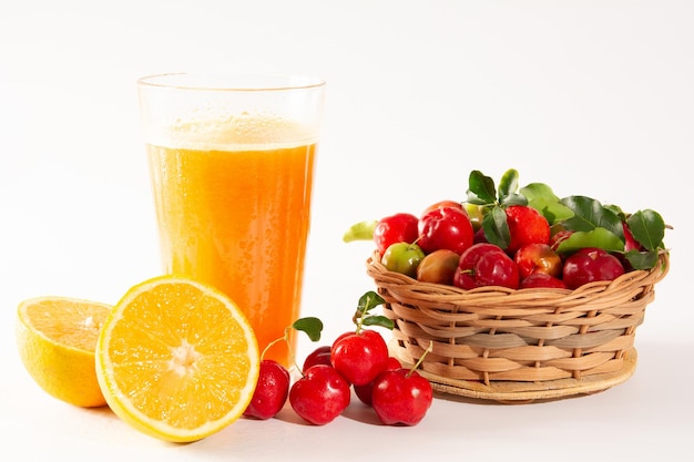 Frischer Bio-Acerola- und Orangensaft in einem Glasbecher mit halben Orangen- und Acerolabeeren in einem Holzkorb auf einem weißen, sauberen Studiofoto in der Vorderansicht