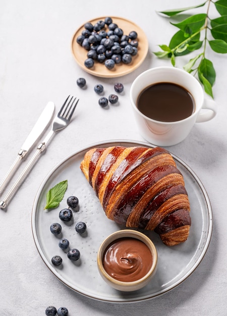 Frischer Croissant mit Schokolade auf einem weißen Teller auf einem hellen Hintergrund mit Blaubeeren und einer Tasse Kaffee Köstliches hausgemachtes Frühstück