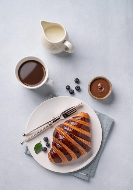 Frischer Croissant mit Schokolade und Blaubeeren auf einem weißen Teller auf einem hellen Hintergrund mit einer Tasse schwarzen Kaffee und einem Milchkrug