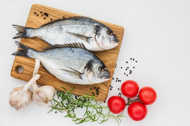 Frischer Dorado-Fisch auf hölzernem Schneidebrett mit Knoblauch und Tomaten auf weißem Tisch. Draufsicht, Kopie