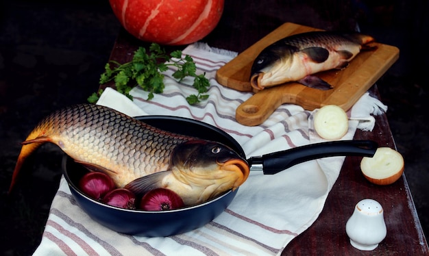 Frischer Fisch auf einer Bratpfanne und auf einem Schneidebrett mit Gemüse auf dunklem Hintergrund Seitenansicht das Konzept der Zubereitung von Fischgerichten mit Gemüse