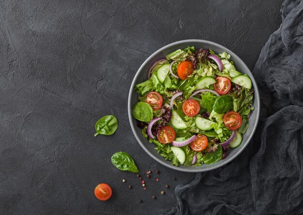 Frischer, gesunder vegetarischer Gemüsesalat mit Tomaten und Gurken, roten Zwiebeln und Spinat in grauer Schüsselplatte auf dunklem Hintergrund mit schwarzer KüchentuchDraufsicht