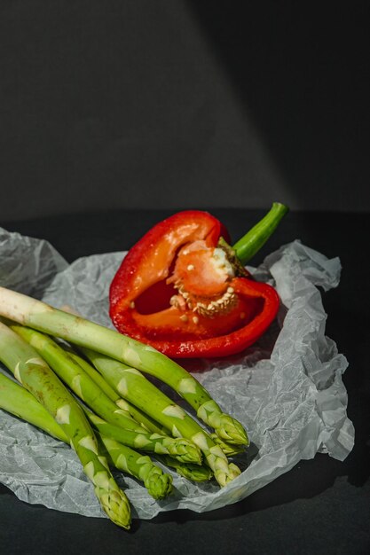 Frischer grüner Spargel und die Hälfte der roten süßen großen Paprika liegen auf weißem Pergament auf dunkelgrauem Hintergrund richtige Ernährung Lifestyle-Platz für Text