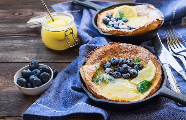 Frischer hausgemachter holländischer Babypfannkuchen mit Zitronenquark und Blaubeeren in Eisenpfannen auf rustikalem Holz.