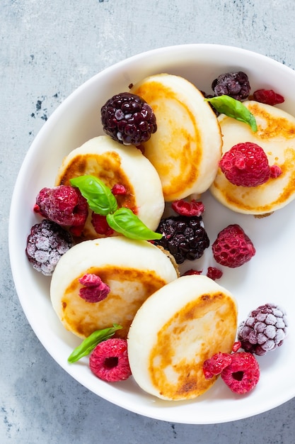 Frischer Hüttenkäse oder Ricotta-Pfannkuchen Syrnikifritters auf einem weißen Teller mit frischen Beeren auf blauem Hintergrund Leckeres gesundes Frühstück Ukrainische russische Küche süße Speisen