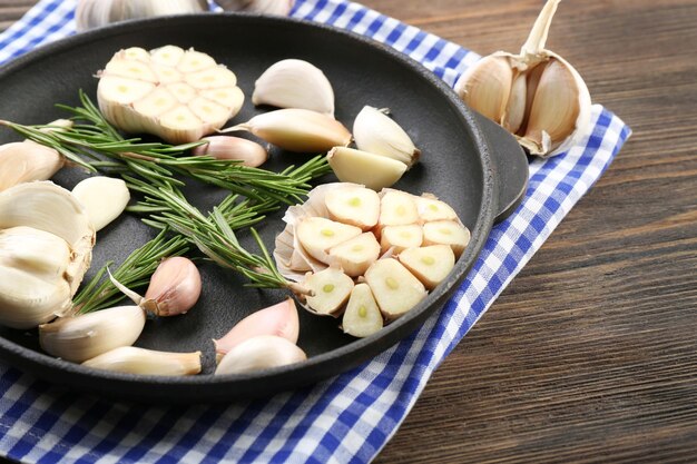 Foto frischer knoblauch mit rosmarin in schwarzer pfanne in der nähe von blau karierter baumwollserviette auf holzhintergrund hautnah