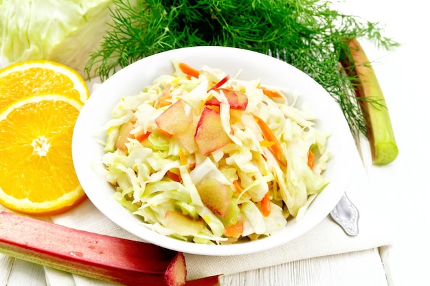 Frischer Kohl-, Karotten- und Rhabarbersalat mit Orangensaft, Honig und Mayonnaise-Dressing in einem Teller auf Serviette, Dill und Gabel auf Holzbretthintergrund