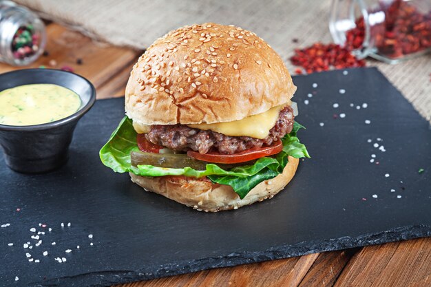 Frischer leckerer Burger mit Käse, Salat, Tomate, Gurke auf schwarzem Stein mit Sauce. Amerikanisches Fast Food. Cheeseburger mit Kopienraum auf hölzernem Hintergrund. Nahaufnahme, selektiver Fokus. Essen. Grillmenü