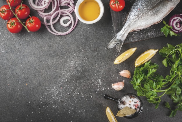 Frischer roher organischer Fisch Dorado, Dorade mit Bestandteilen für das Kochen der Zitrone, der Tomaten, der Zwiebel, des Salzes, des Pfeffers, des Lorbeers, der Grüns, des Öls. Auf altem hölzernem Schneidebrett auf schwarzem Steintischplatteansicht-Kopienraum