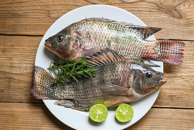 Frischer roher Tilapia-Fisch aus der Tilapia-Farm Tilapia mit weißer Platte mit Rosmarin-Zitronen-Limette auf Holzhintergrund