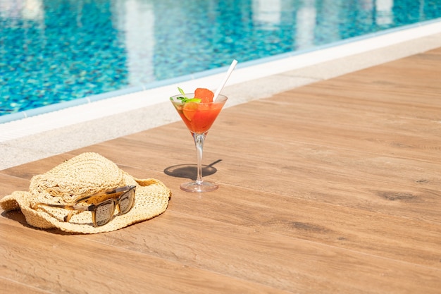 Frischer roter Cocktail mit Eis im Glas, Strandhut und Sonnenbrille am Swimmingpool. Tropischer Saft im Luxusurlaub. Konzept Sommerurlaub und Reisen. Foto in hoher Qualität