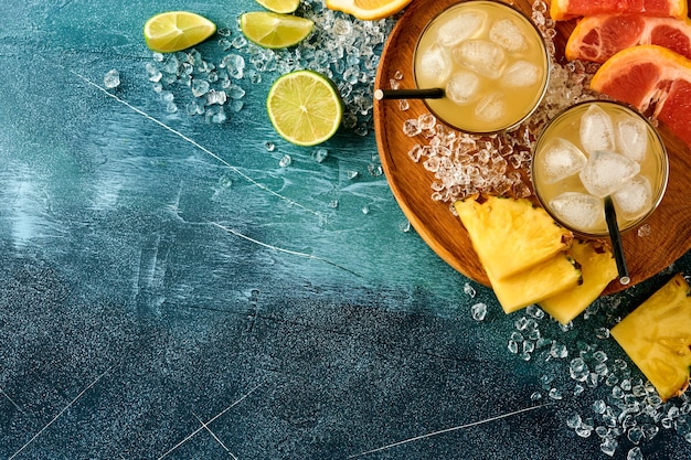 Frischer Saft Ananas mit Eis und Vielfalt in Scheiben geschnitten reife tropische Früchte Grapefruit, Orange, Limette, Zitrone, flache Zusammensetzung, auf dunkelblauem Steinhintergrund.