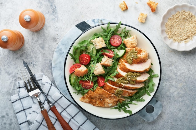 Frischer Salat mit gegrilltem Hähnchenfilet, Rucola, Tomaten, Gurke und Pesto-Sauce, Sesamsamen mit Olivenöl in weißer Schüssel auf hellem Schieferhintergrund. Gesundes Mittagsmenü