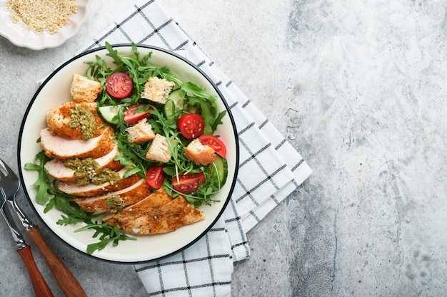 Frischer Salat mit gegrilltem Hähnchenfilet, Rucola, Tomaten, Gurke und Pesto-Sauce, Sesamsamen mit Olivenöl in weißer Schüssel auf hellem Schieferhintergrund. Gesundes Mittagsmenü