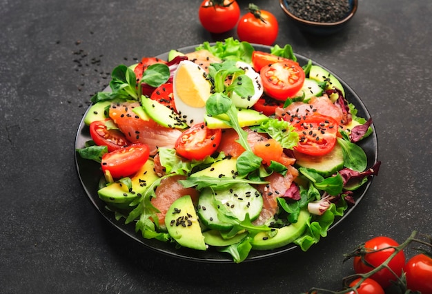 Frischer Salat mit gesalzenem Lachs, Avocado, Ei, Sesamsamen, Olivenöl, Tomaten und gemischten Kräutern Keto-Diät-Essen, schwarzer Hintergrund, Draufsicht